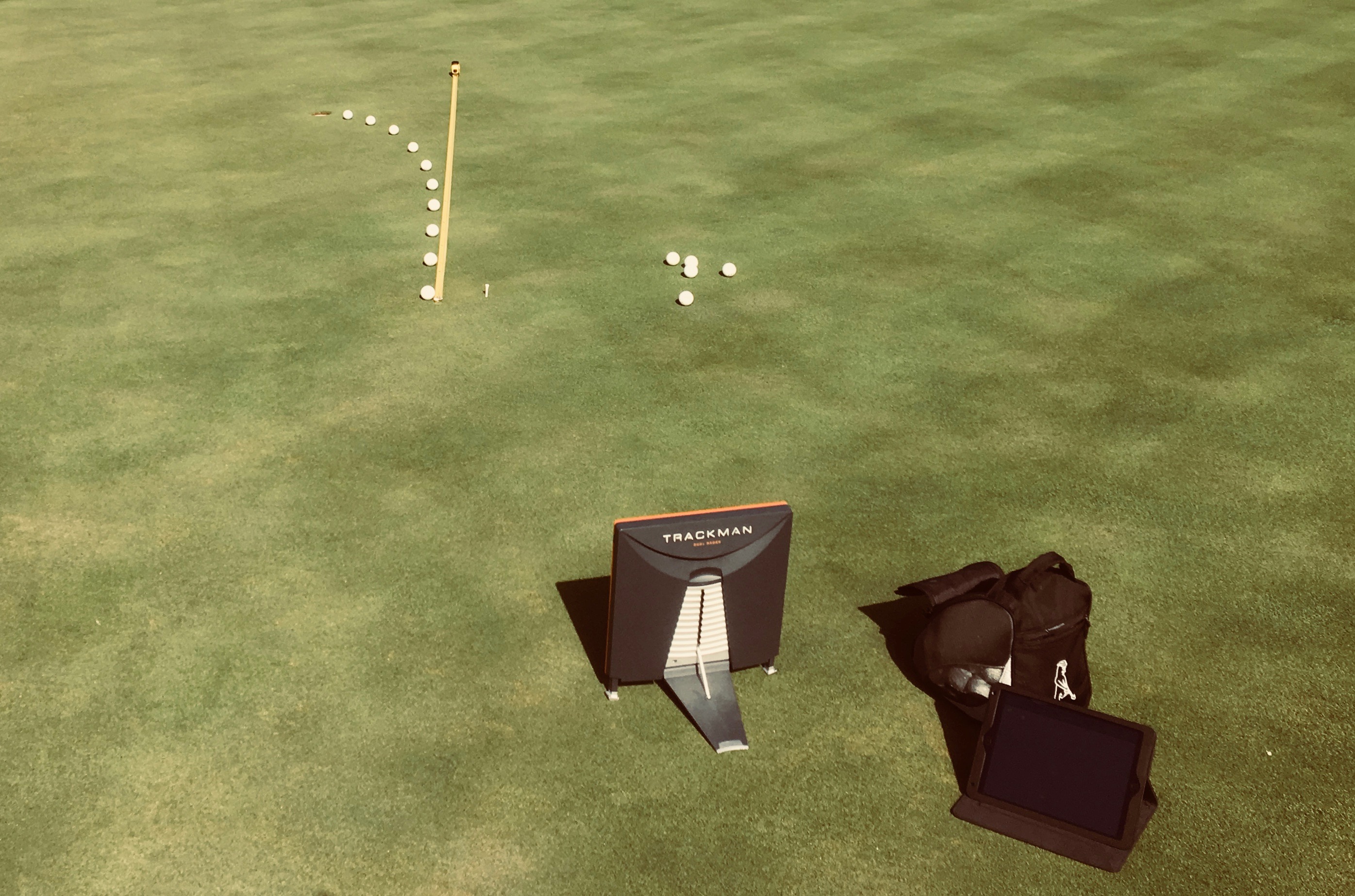 TrackMan Putt on a practice green, with a line of balls tracing the curve of a breaking putt into the hole beside a straight alignment stick
